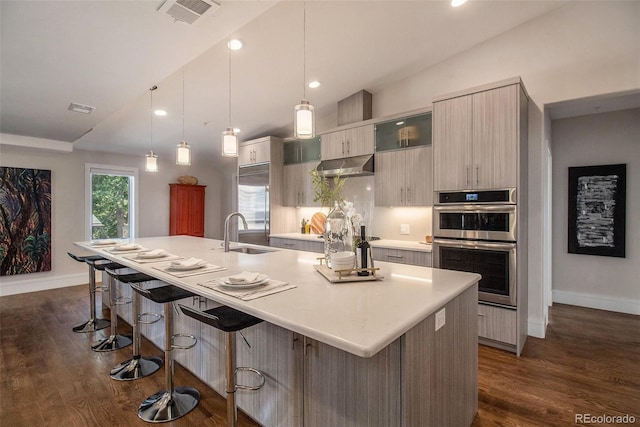 kitchen with sink, stainless steel double oven, hanging light fixtures, an island with sink, and vaulted ceiling