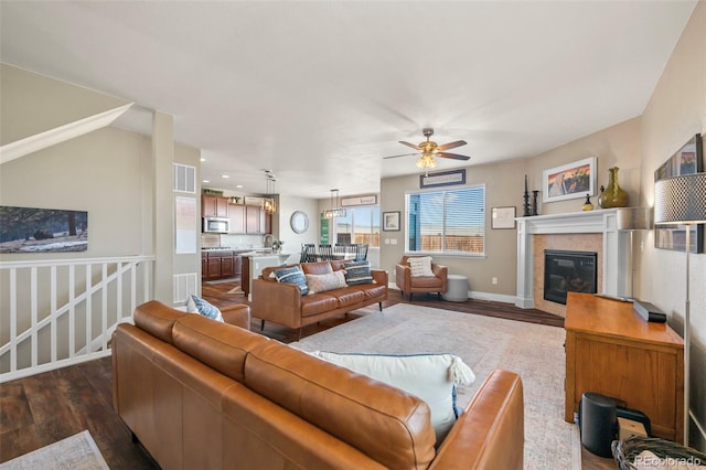 living room featuring ceiling fan, a tile fireplace, visible vents, baseboards, and dark wood-style floors