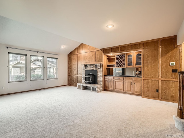 unfurnished living room with a wood stove, vaulted ceiling, light colored carpet, wooden walls, and baseboards