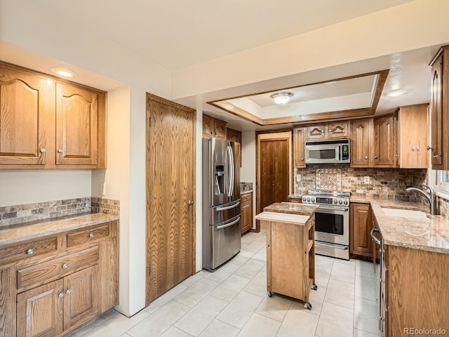 kitchen with appliances with stainless steel finishes, a sink, a tray ceiling, backsplash, and brown cabinetry