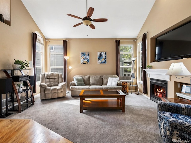 carpeted living room with vaulted ceiling, a ceiling fan, a warm lit fireplace, baseboards, and wood finished floors