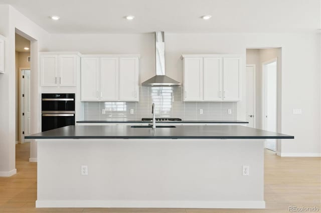 kitchen featuring wall chimney exhaust hood, sink, double oven, a kitchen island with sink, and white cabinets