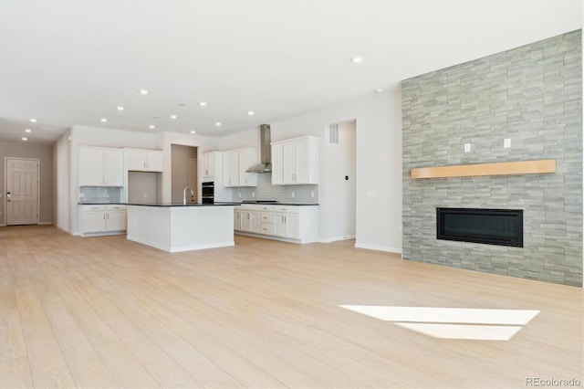 kitchen with wall chimney range hood, a kitchen island with sink, white cabinetry, a large fireplace, and decorative backsplash