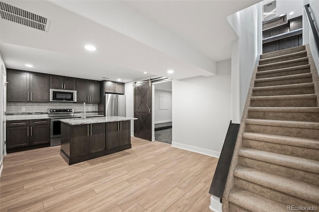 kitchen featuring appliances with stainless steel finishes, backsplash, a barn door, a center island with sink, and light hardwood / wood-style floors