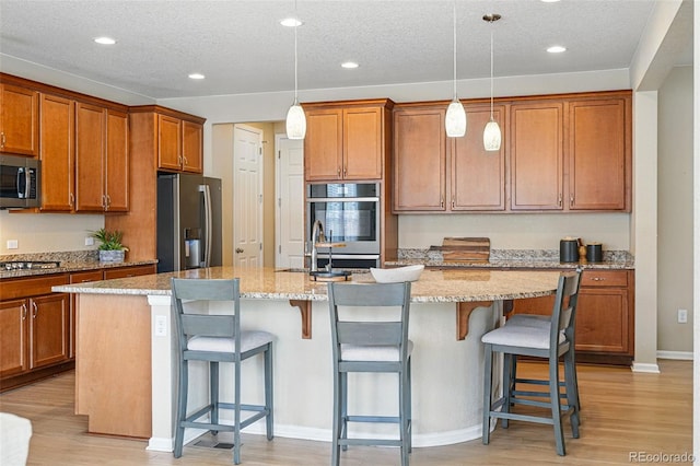 12623 Canoe St, Firestone, CO 80504 - kitchen with stainless steel appliances, a kitchen island with sink, brown cabinetry, light wood-style flooring, and recessed lighting