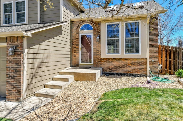 doorway to property featuring brick siding, fence, and a shingled roof