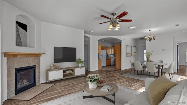 living room featuring ceiling fan with notable chandelier, baseboards, a tiled fireplace, and wood finished floors