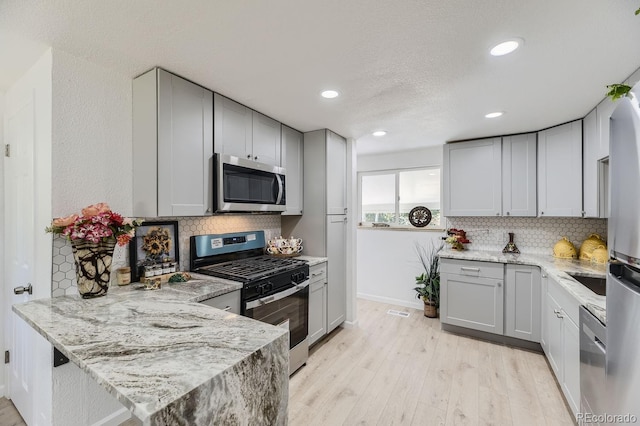 kitchen with light wood-style flooring, gray cabinetry, appliances with stainless steel finishes, light stone countertops, and tasteful backsplash