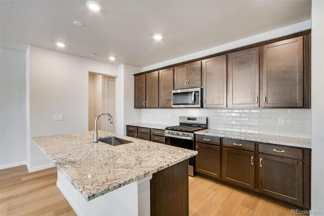 kitchen featuring an island with sink, sink, dark brown cabinetry, stainless steel appliances, and light stone countertops