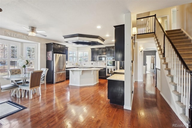 kitchen with appliances with stainless steel finishes, backsplash, a center island, dark wood-type flooring, and ceiling fan