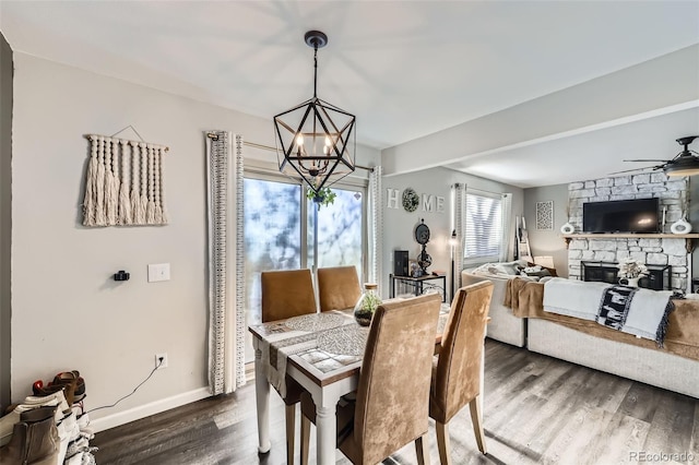 dining space with ceiling fan with notable chandelier, dark wood-type flooring, and a stone fireplace