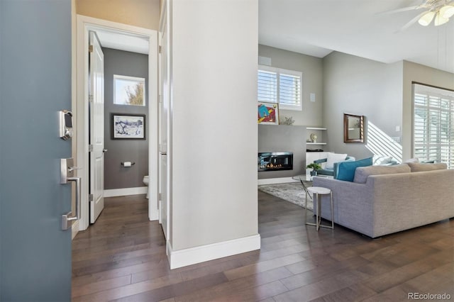 interior space featuring dark wood-style flooring, a glass covered fireplace, a ceiling fan, and baseboards