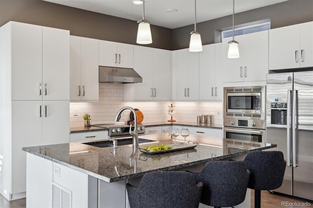 kitchen with under cabinet range hood, stainless steel appliances, hanging light fixtures, decorative backsplash, and a kitchen bar