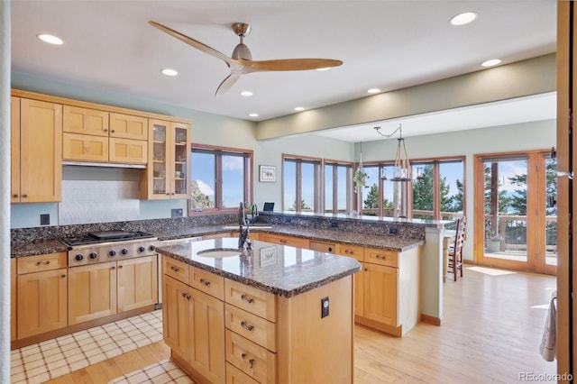 kitchen with stainless steel gas stovetop, dark stone counters, a center island with sink, ceiling fan with notable chandelier, and sink