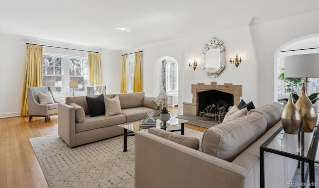 living room featuring arched walkways, a fireplace with raised hearth, ornamental molding, light wood-type flooring, and baseboards