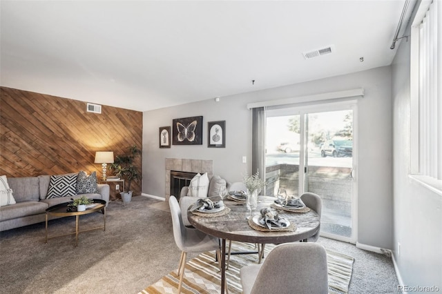 dining space with light colored carpet, visible vents, an accent wall, wooden walls, and a tile fireplace