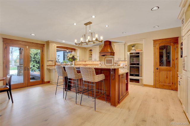 kitchen featuring hanging light fixtures, stainless steel double oven, an island with sink, cream cabinetry, and custom exhaust hood