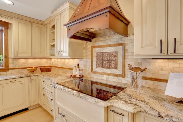 kitchen featuring premium range hood, cream cabinets, black electric stovetop, light stone countertops, and tasteful backsplash