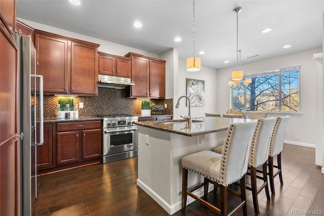 kitchen featuring decorative light fixtures, an island with sink, sink, dark stone counters, and high end appliances