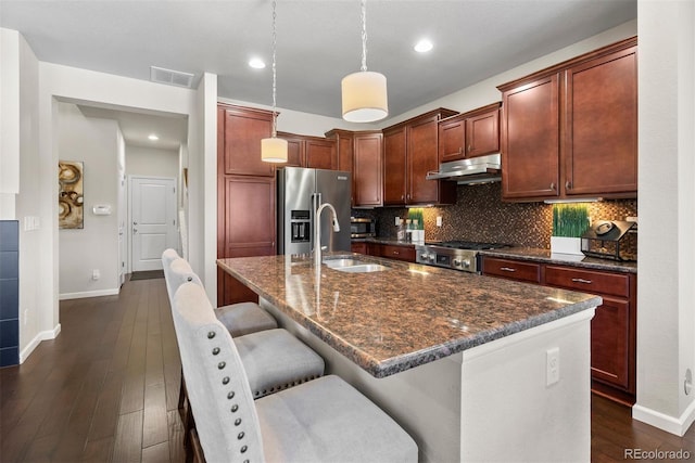 kitchen with high end fridge, stove, a center island with sink, decorative light fixtures, and dark stone counters