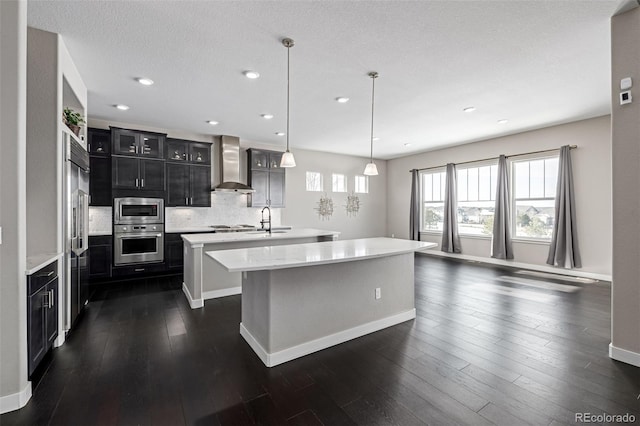 kitchen with wall chimney range hood, stainless steel appliances, tasteful backsplash, an island with sink, and decorative light fixtures