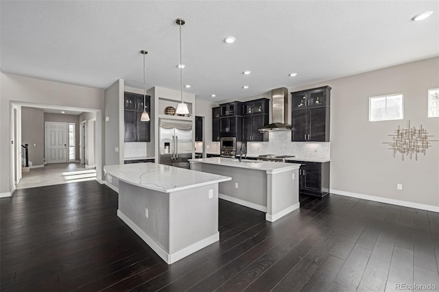 kitchen featuring decorative light fixtures, backsplash, a kitchen island with sink, built in appliances, and wall chimney exhaust hood