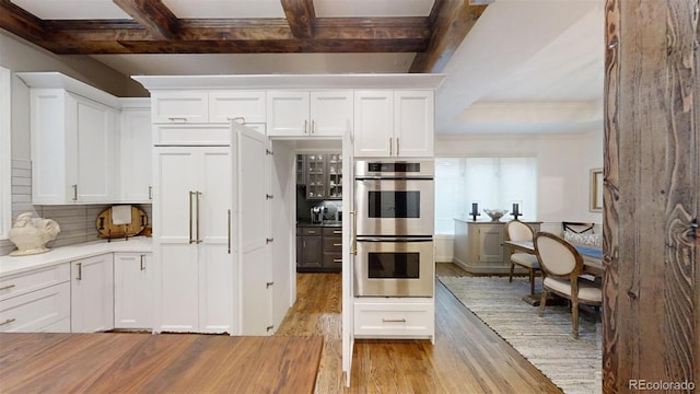 kitchen with light wood finished floors, tasteful backsplash, stainless steel double oven, white cabinetry, and beam ceiling