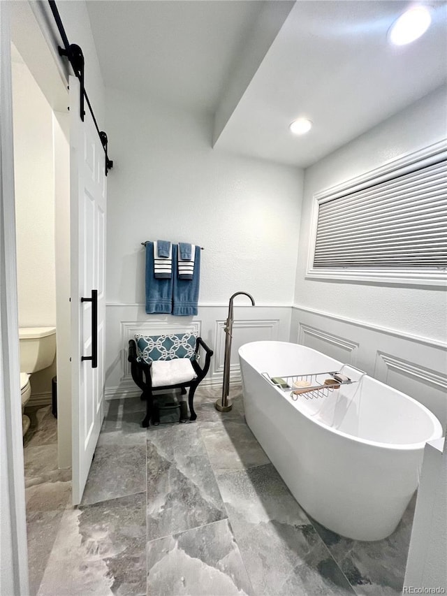 bathroom featuring toilet, a soaking tub, a decorative wall, and wainscoting