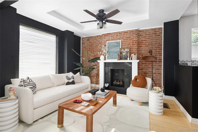 living room with brick wall, wood-type flooring, a tray ceiling, ceiling fan, and a premium fireplace