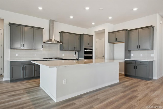 kitchen featuring light wood finished floors, a kitchen island with sink, a sink, double oven, and wall chimney exhaust hood