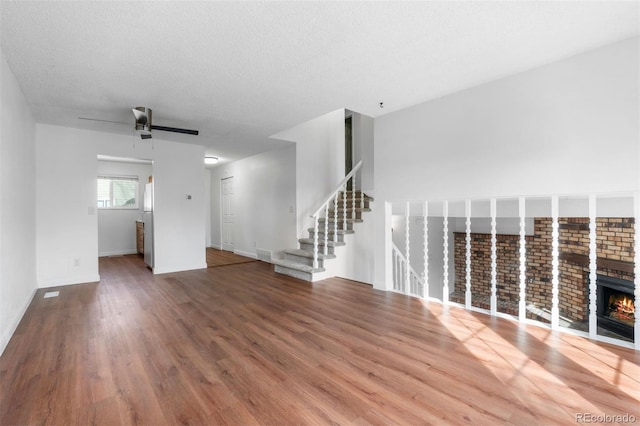 unfurnished living room featuring ceiling fan, stairway, wood finished floors, a textured ceiling, and a brick fireplace