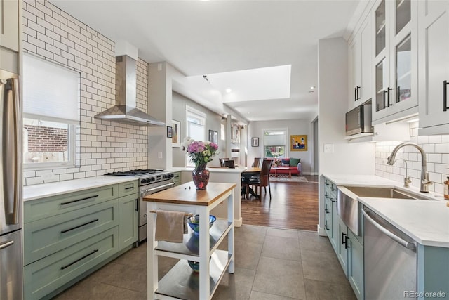 kitchen featuring wall chimney range hood, light countertops, appliances with stainless steel finishes, green cabinetry, and a sink