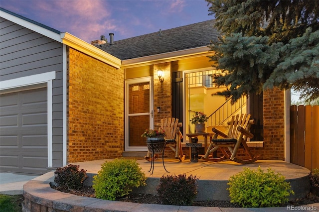 exterior entry at dusk featuring brick siding, fence, an attached garage, and a shingled roof
