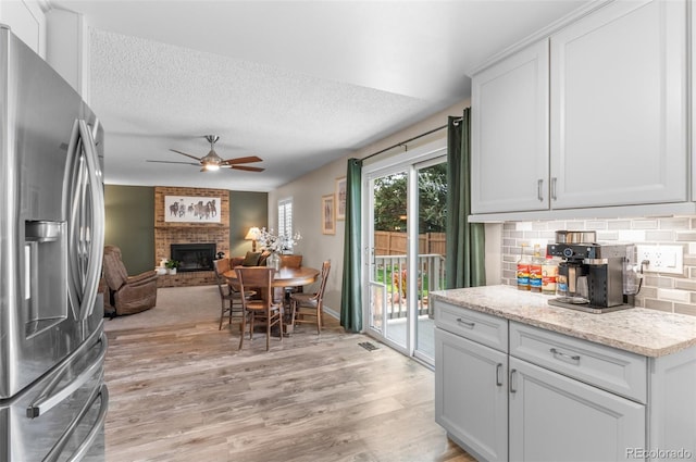 kitchen with stainless steel fridge with ice dispenser, a ceiling fan, a brick fireplace, light wood finished floors, and backsplash