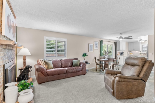 living area featuring a brick fireplace, a ceiling fan, baseboards, a textured ceiling, and light colored carpet