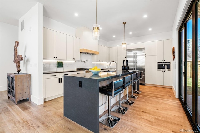 kitchen with dark countertops, a center island with sink, stainless steel appliances, a sink, and white cabinetry