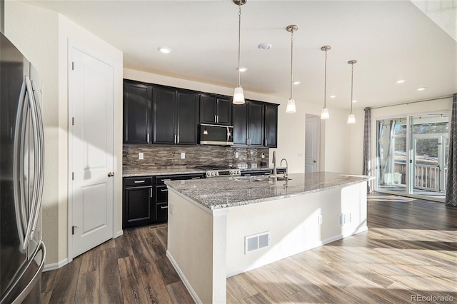 kitchen featuring dark wood-type flooring, stainless steel appliances, tasteful backsplash, decorative light fixtures, and a kitchen island with sink