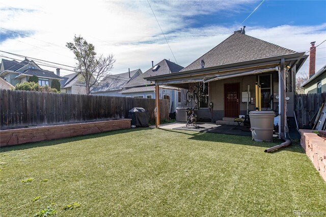rear view of house with a lawn, a patio area, cooling unit, a fenced backyard, and a shingled roof