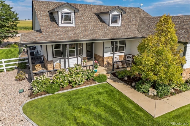 new england style home with fence, covered porch, a shingled roof, a front lawn, and stone siding