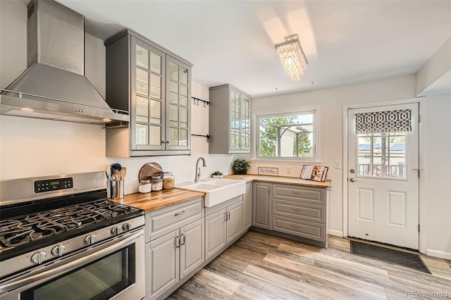 kitchen featuring stainless steel gas range oven, wall chimney exhaust hood, a sink, gray cabinetry, and butcher block counters