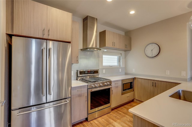 kitchen featuring light brown cabinets, wall chimney exhaust hood, light hardwood / wood-style flooring, appliances with stainless steel finishes, and tasteful backsplash