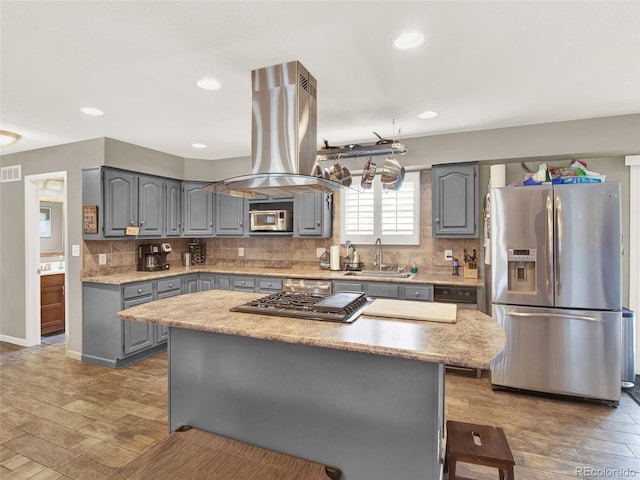 kitchen with decorative backsplash, appliances with stainless steel finishes, a sink, island range hood, and gray cabinets