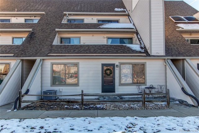 snow covered property entrance with cooling unit, roof with shingles, and fence