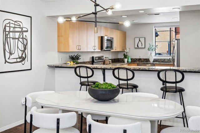 kitchen featuring stainless steel appliances, light brown cabinetry, a breakfast bar, dark stone countertops, and a peninsula