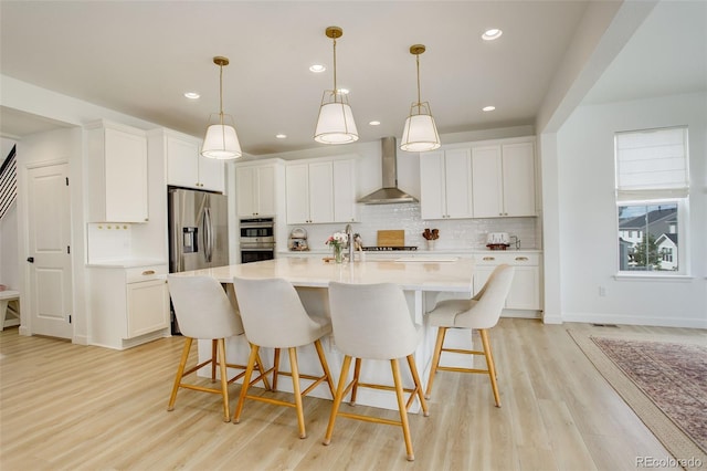kitchen featuring wall chimney exhaust hood, appliances with stainless steel finishes, decorative backsplash, light countertops, and light wood-style floors