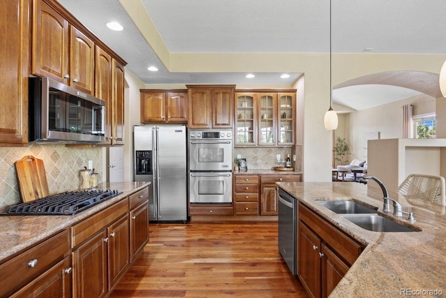 7472 Iridium Way, Castle Rock, CO 80108 - kitchen featuring appliances with stainless steel finishes, arched walkways, light stone counters, light wood-style flooring, and glass insert cabinets