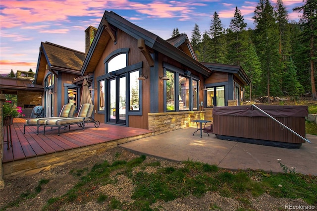 back of property at dusk featuring stone siding, french doors, a wooden deck, a chimney, and a hot tub