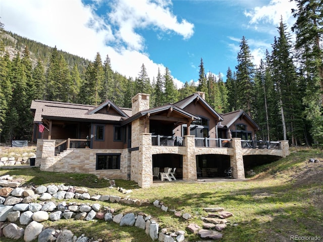 back of house featuring a forest view, stone siding, a chimney, and a patio area