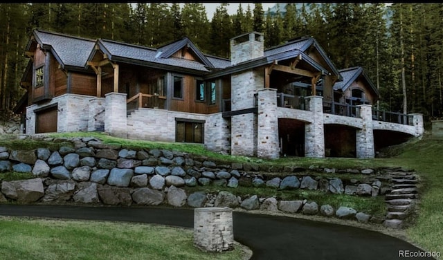 rear view of property with a chimney, stone siding, an attached garage, and a view of trees