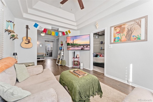 living room with dark wood-type flooring, a raised ceiling, a ceiling fan, and baseboards